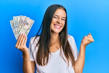 Young hispanic woman holding dollars screaming proud, celebrating victory and success very excited...