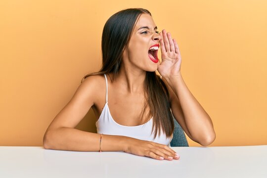 Young hispanic woman wearing casual clothes sitting on the table shouting and screaming loud to side with hand on mouth. communication concept.