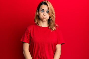 Hispanic young woman wearing casual red t shirt puffing cheeks with funny face. mouth inflated with air, crazy expression.