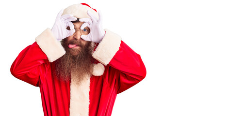 Handsome young red head man with long beard wearing santa claus costume doing ok gesture like binoculars sticking tongue out, eyes looking through fingers. crazy expression.