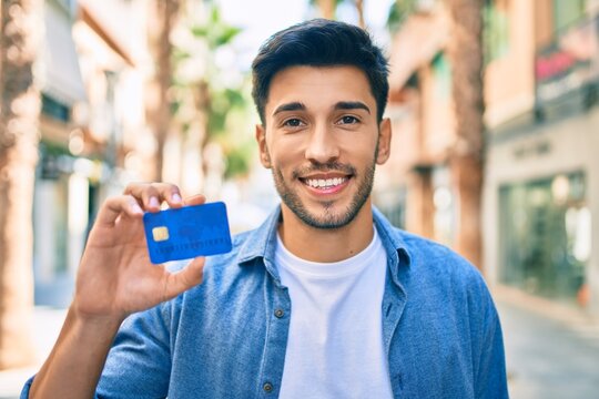 Young latin man smiling happy holding credit card walking at the city.