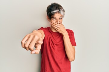 Young hispanic man wearing casual clothes laughing at you, pointing finger to the camera with hand over mouth, shame expression