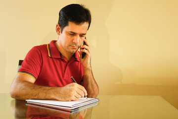 Adult man sitting at desk in his living room, with yellow background wall, talking on cell phone and writing notes on paper