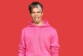 Young hispanic man wearing casual pink sweatshirt sticking tongue out happy with funny expression. emotion concept.