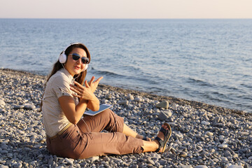 Young woman in white headphones and dark glasses sits on the seashore, moves hand gestures to the rhythm of the music
