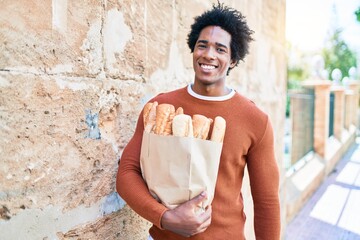 Naklejka na ściany i meble Young handsome african american man smiling happy. Standing with smile on face holding paper bag with bread walking at town street.