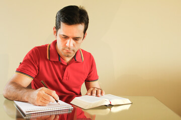 Adult man sitting at the table in his living room, with yellow background wall, reading the bible and writing notes on a paper