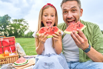Portrait of happy dad and his adorable little daughter looking at camera while eating watermelon,...