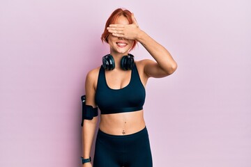 Obraz premium Young beautiful redhead woman wearing gym clothes and using headphones smiling and laughing with hand on face covering eyes for surprise. blind concept.