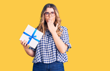 Young caucasian woman holding gift covering mouth with hand, shocked and afraid for mistake. surprised expression