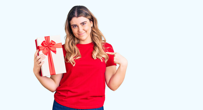 Young caucasian woman holding gift pointing finger to one self smiling happy and proud