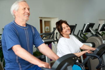 Fototapeta na wymiar Senior couple spinning on fitness bikes. Happy elderly woman training on machine at gym.