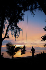Dark silhouette of a man swinging a woman on a swing at a beautiful tropical sunset