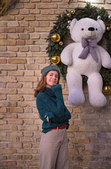 Stylish young woman in knitted sweater and a hat near the Christmas street decorations on the brick wall