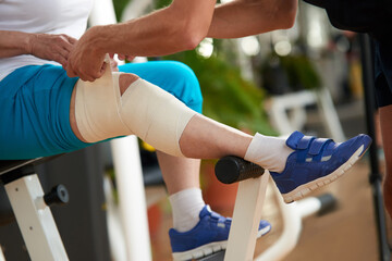 Male hands wrapping bandage around knee. Unrecognizable mature woman with injured leg at gym. Sport and healthcare theme.