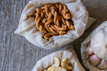 Variety of different groceries in cotton bags packed for storage: pasta, nuts, garic on stone background. Eco-friendly organic bio kitchen products. Zero waste sustainable plastic free lifestyle