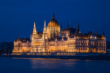 Fototapeta na wymiar A spectacularly illuminated grandiose neo-Gothic building on the river embankment in a European city. Blue hour, clear dark blue sky. No people