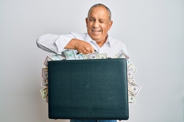 Handsome senior man holding briefcase full of dollars winking looking at the camera with sexy...