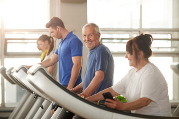 Old happy man runs on treadmill. People training at modern gym. Regular sports trainings help to...
