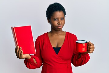 Young african woman with short hair reading a book and drinking a cup of coffee skeptic and...
