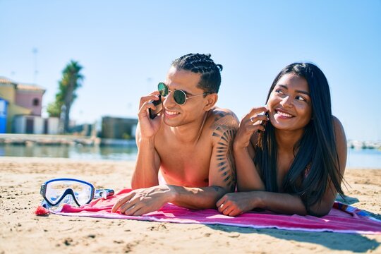 Young latin couple talking on the smartphone lying on the sand at the beach.