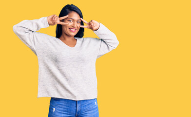 Fototapeta premium Hispanic woman with long hair wearing casual clothes doing peace symbol with fingers over face, smiling cheerful showing victory