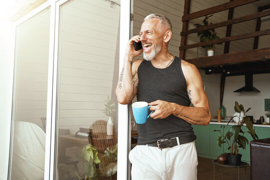 Enjoying good talk. Happy middle aged caucasian man talking by mobile phone and laughing while drinking coffee near balcony of his modern flat