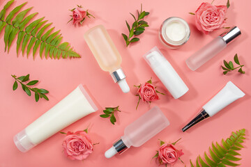 Flat lay cosmetic bottles and containers with roses and green fern leaves on pink background