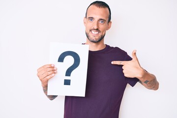 Young handsome man with tattoo holding question mark pointing finger to one self smiling happy and proud