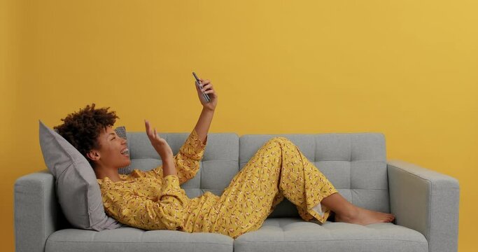 Happy relaxed young Afro American woman lying on sofa in nightwear makes video call waves hello to interlocutor uses free internet connection at home rests from work poses against yellow wall
