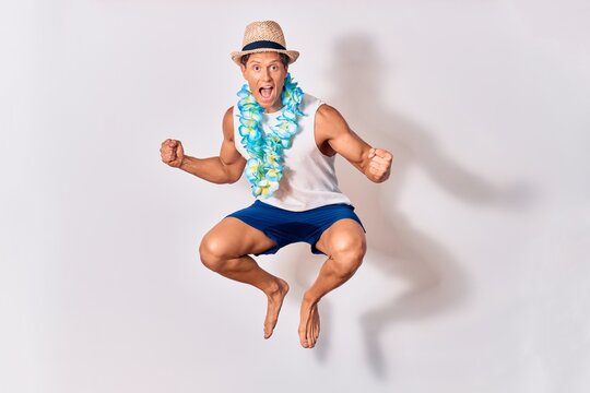 Young handsome hispanic tourist man on vacation wearing summer clothes and hawaiian lei smiling happy. Jumping with smile on face celebrating with fists up over isolated white background