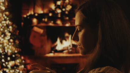 A beautiful girl is sitting in a cozy interior. Dreaming, thinking. Festive interior with lights, Christmas tree and fireplace in the background. Winter evening, cozy. Soft focus, depth of field