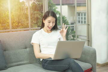 Naklejka premium Freelance Jobs workation concept.Positive smile asian girl sitting on sofa with laptop and showing ok sign.