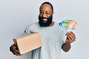 Young african american man holding delivery box and swiss franc banknotes winking looking at the...
