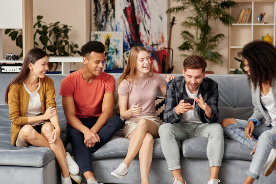 All you need is friends. Group of happy young multicultural people in casual wear looking at smartphone, enjoying time together while sitting on the sofa in the living room