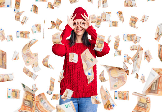 Young beautiful girl wearing sweater and wool cap doing ok gesture like binoculars sticking tongue out, eyes looking through fingers. crazy expression.