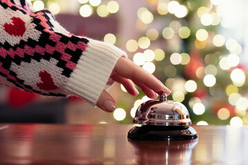Obraz na płótnie Canvas Hand of guest ringing reception bell on desk of guesthouse, hotel at christmas time. Color shining garland on christmas tree on background. Travel concept.