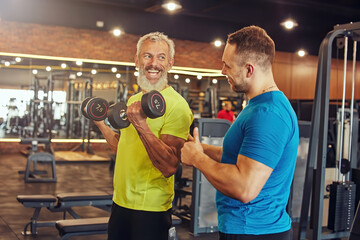 Positive mature man in sportswear doing weight exercises with his young personal trainer, working out at gym with fitness instructor