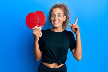 Beautiful caucasian teenager girl holding red ping pong rackets surprised with an idea or question pointing finger with happy face, number one