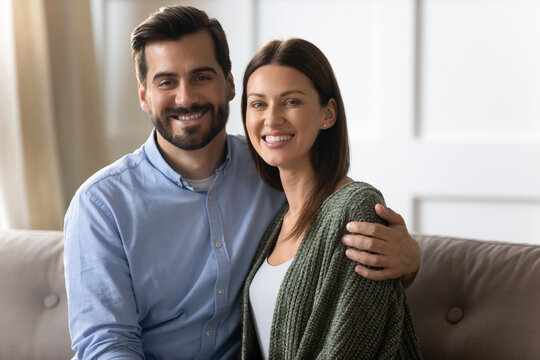 Headshot portrait of happy young Caucasian couple sit on couch at home hug cuddle show love and care in relations. Profile picture of smiling man and woman spouses enjoy good family relationship.