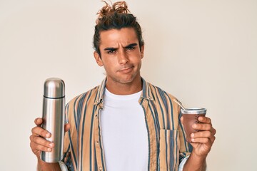 Young hispanic man drinking mate infusion clueless and confused expression. doubt concept.
