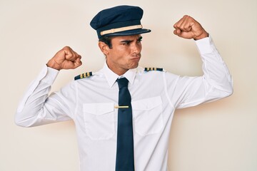 Young hispanic man wearing airplane pilot uniform showing arms muscles smiling proud. fitness...