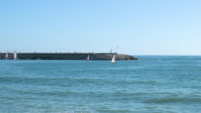 Sailing boats sailing near the harbor and people strolling away on the jetty.