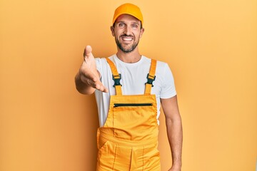Young handsome man wearing handyman uniform over yellow background smiling friendly offering handshake as greeting and welcoming. successful business.