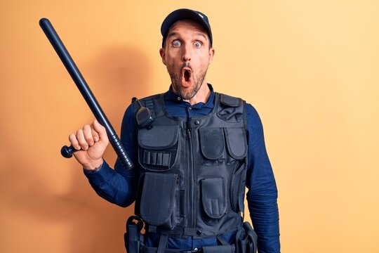 Handsome policeman wearing uniform and bulletprof holding baton over yellow background scared and amazed with open mouth for surprise, disbelief face