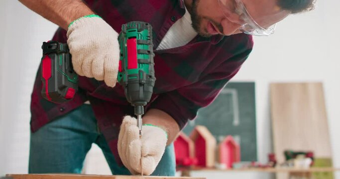 Concentrated young bearded carpenter wearing jeans and a plaid flannel shirt drilling a hole in a piece of wood with a portable electric drill in his carpentry workshop.
