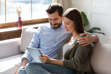 Young Caucasian couple sit relax on couch at home look at tablet screen browsing wireless internet on gadget. Happy man and woman renters rest on sofa in living room use modern pad device together.