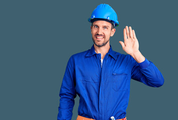 Young handsome man wearing worker uniform and hardhat waiving saying hello happy and smiling, friendly welcome gesture