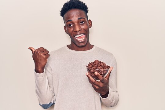 Young african american man holding chocolate cereals pointing thumb up to the side smiling happy with open mouth
