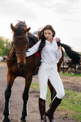 Obraz premium A young pretty girl rider poses near a thoroughbred stallion on a ranch. Horse riding, horse racing
